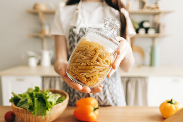 Woman in apron holding glass jar with wheat pasta in modern kitchen.