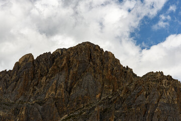 Close-up photo of a mountain peak in the French Alps, dark rock and clouds, blue sky. At the top very small mountaineers can be seen,