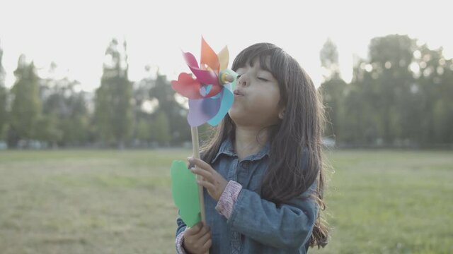 Adorable Latin Girl Blowing On Paper Fan Outdoors. Cute Brunette Child Standing And Playing With Colorful Toy On Wooden Stick In City Park. Static Camera. Childhood, Weekend And Summer Concept