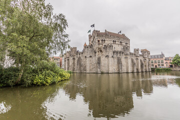 Fototapeta na wymiar Panoramic view of calm waters surrounding the medieval castle of Gravensteen (Castle of the Counts), reflection in the water surface and green vegetation, cloudy day with a gray sky in Ghent, Belgium