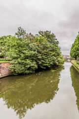 Fototapeta na wymiar Canal with calm waters and reflection on the water surface, lush trees with green foliage and a bridge in the background, cloudy day with a sky covered by dense clouds in the city of Ghent, Belgium