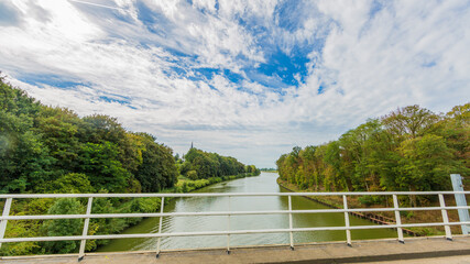 Fototapeta na wymiar Juliana canal surrounded by lush trees seen from the Elsloo bridge with its white metal railing, sunny day with a blue sky and white clouds in South Limburg, the Netherlands Holland