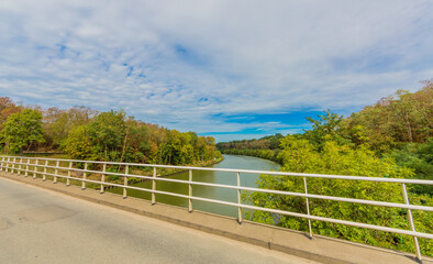 Fototapeta premium Elsloo vehicular bridge with its white metal railing, Juliana canal surrounded by greenery and lush trees with green foliage, sunny day with a blue sky and white clouds in South Limburg, Netherlands