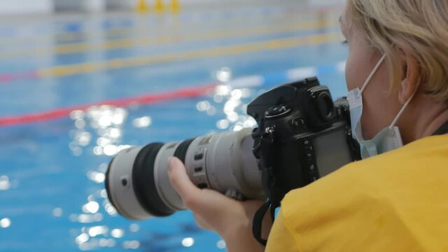 a sports photographer works in a pool wearing a face mask to prevent the spread of covid 19. Blond photographer in mask. Shoots with DSLR and telephoto lens