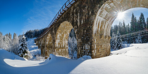 Stone viaduct (arch bridge) on railway through mountain snowy fir forest. Snow drifts  on wayside...