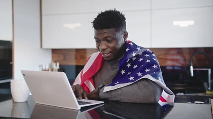 African american man with USA flag over his shoulders watching game on his laptop with cup of tea on the table. Surprised wace expression. High quality 4k footage - Powered by Adobe