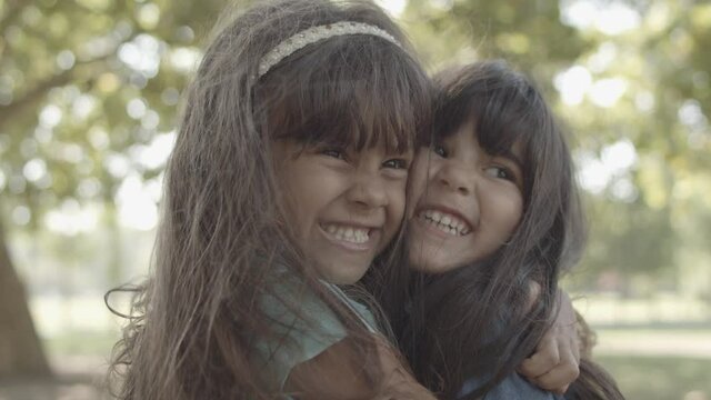 Closeup portrait of Latin girls hugging each other and smiling. Adorable happy little sisters standing in city park, laughing, embracing and kissing. Static camera. Childhood and friendship concept