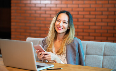 A Korean girl works with pleasure at a laptop and a mobile phone in a loft with brick walls. Modern business running in an urban lifestyle. Work in multitasking mode.