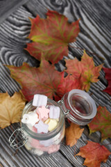 Marshmallows of different colors. They lie in a glass jar. Autumn maple leaves are scattered around.