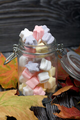 Marshmallows of different colors. They lie in a glass jar. Autumn maple leaves are scattered around.