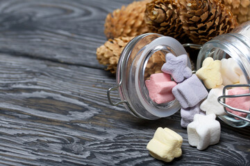 Marshmallows of different colors. They lie in an overturned glass jar. Nearby are spruce cones. Against the background of black pine boards.