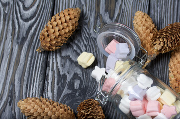Marshmallows of different colors. They lie in an overturned glass jar. Nearby are spruce cones. Against the background of black pine boards.