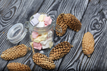 Marshmallows of different colors. They lie in  glass jar. Nearby are spruce cones. Against the background of black pine boards.