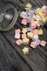 Marshmallows of different colors. They lie in an overturned glass jar. Against the background of black pine boards.