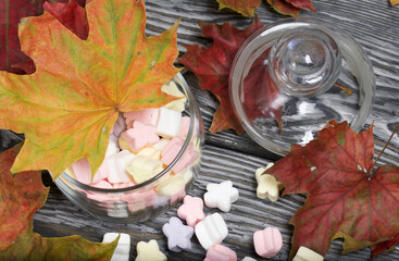 Marshmallows of different colors. They lie in a glass jar. Autumn maple leaves are scattered around.