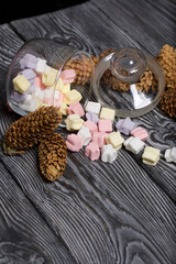 Marshmallows of different colors. They lie in an overturned glass jar. Nearby are spruce cones. Against the background of black pine boards.