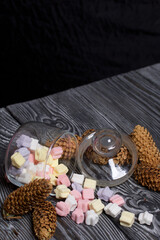 Marshmallows of different colors. They lie in an overturned glass jar. Nearby are spruce cones. Against the background of black pine boards.