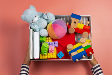 Donation concept. Kid hands with donate box with clothes, books and toys on pink background. Top view