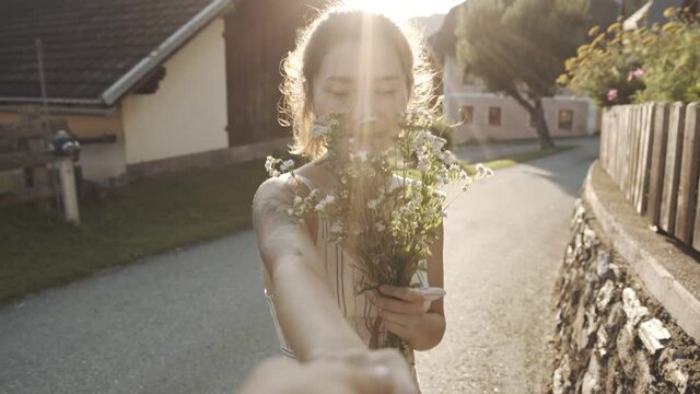 Follow me, rear view of young asian girl with bouquet of flowers guiding guy along village, looking at camera. First-person view of woman walking at sunset, inviting hand to join. Lifestyle concept.