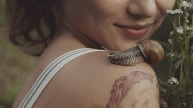 Woman holding bouquet of wild flowers or daisies in her hands. Close-up of snail slowly crawling along shoulder of young girl. Snail leaves muscus on skin. Concept of nature, cosmetology, spa, beauty.