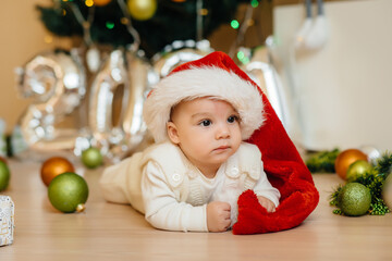 Cute smiling baby is lying under a festive Christmas tree and playing with gifts. Christmas and New Year celebrations