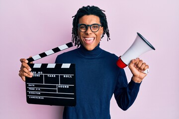 Young african american man holding video film clapboard and megaphone smiling with a happy and cool...