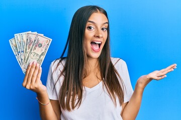 Young hispanic woman holding dollars celebrating achievement with happy smile and winner expression with raised hand