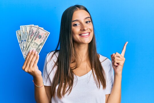 Young hispanic woman holding dollars smiling happy pointing with hand and finger to the side