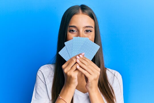 Young hispanic woman playing gambling poker covering face with cards smiling with a happy and cool smile on face. showing teeth.