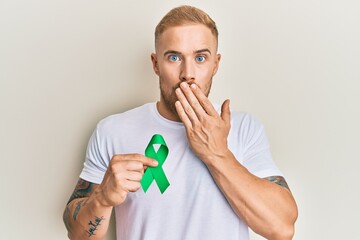 Young caucasian man holding support green ribbon covering mouth with hand, shocked and afraid for...