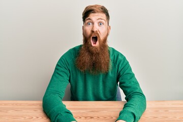 Young irish redhead man wearing casual clothes sitting on the table afraid and shocked with...