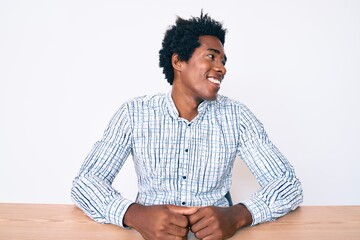 Handsome african american man with afro hair wearing casual clothes sitting on the table looking away to side with smile on face, natural expression. laughing confident.