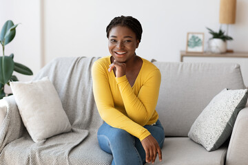 Positive young black woman posing on sofa at home, smiling at camera