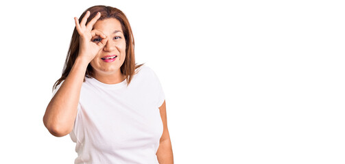 Middle age latin woman wearing casual white tshirt doing ok gesture with hand smiling, eye looking through fingers with happy face.