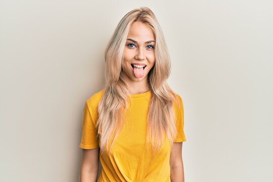 Beautiful caucasian blonde girl wearing casual tshirt sticking tongue out happy with funny expression. emotion concept.