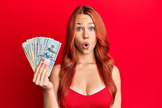 Young beautiful redhead woman holding dollars scared and amazed with open mouth for surprise, disbelief face