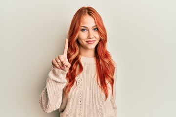 Young beautiful redhead woman wearing casual winter sweater showing and pointing up with finger number one while smiling confident and happy.