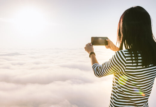 Brunette woman holds smartphone and takes a photo of the sunrise above the clouds. Concept of solo travel joy and memories.