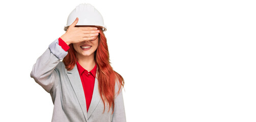 Young redhead woman wearing architect hardhat smiling and laughing with hand on face covering eyes for surprise. blind concept.