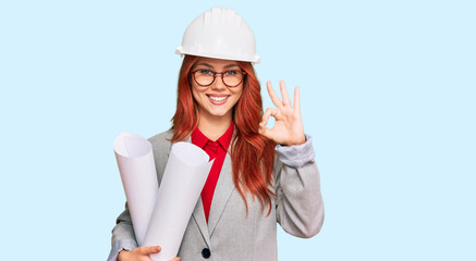 Young redhead architect woman wearing safety helmet holding blueprints doing ok sign with fingers, smiling friendly gesturing excellent symbol