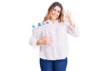 Young caucasian woman holding recycling plastic bottles doing ok sign with fingers, smiling friendly gesturing excellent symbol