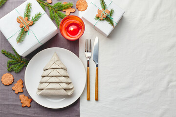 Napkin in the form of a Christmas tree on a plate on white tablecloth with gifts and decorations with fir sprigs and gingerbread cookies