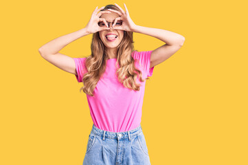 Young beautiful caucasian woman with blond hair wearing casual clothes and glasses doing ok gesture like binoculars sticking tongue out, eyes looking through fingers. crazy expression.