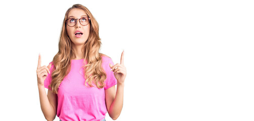 Fototapeta na wymiar Young beautiful caucasian woman with blond hair wearing casual clothes and glasses amazed and surprised looking up and pointing with fingers and raised arms.
