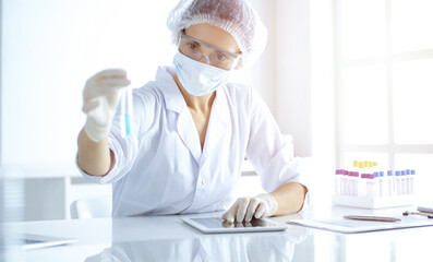 Professional female scientist in protective eyeglasses researching tube with reagents in sunny laboratory. Medicine and science researching