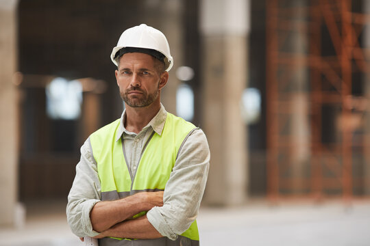 Waist up portrait of mature construction worker looking at camera while standing with arms crossed at construction site, copy space