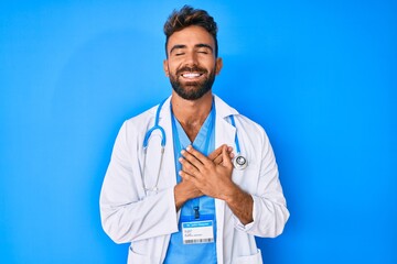 Young hispanic man wearing doctor uniform and stethoscope smiling with hands on chest with closed...
