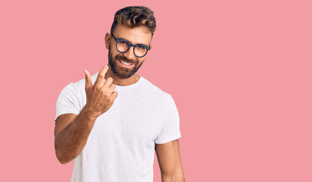 Young hispanic man wearing casual clothes and glasses beckoning come here gesture with hand inviting welcoming happy and smiling