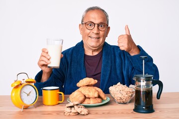 Senior handsome man with gray hair sitting on the table drinking a glass milk for breakfast smiling happy and positive, thumb up doing excellent and approval sign