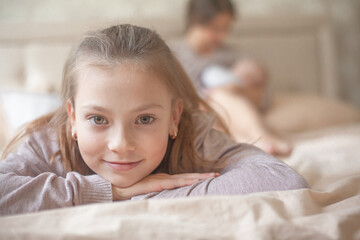 Portrait of preteen girl lies on the bed, while mother take care the newborn baby on the background